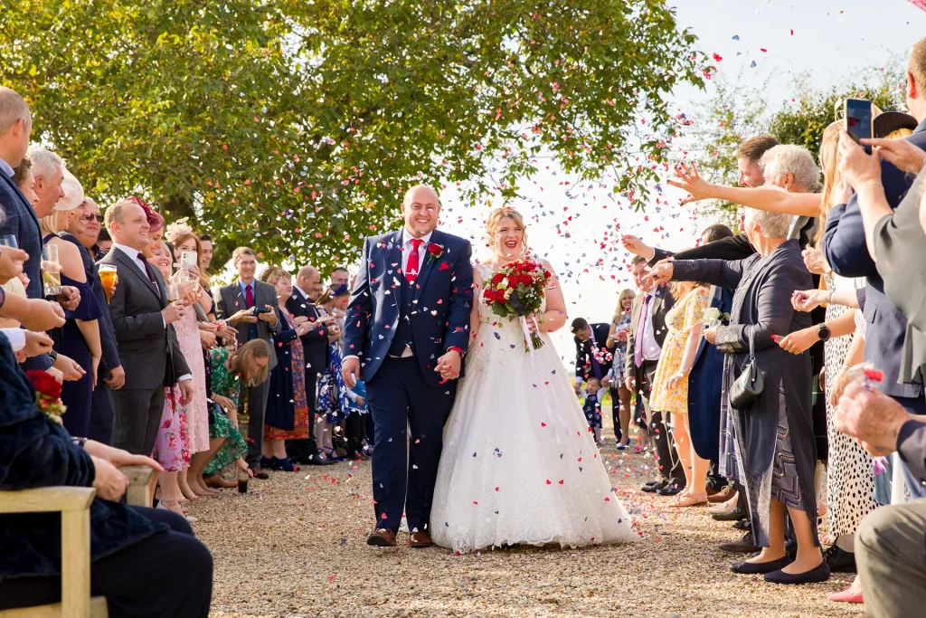 A joyful bride in a white gown and groom in a navy suit walk down an aisle outdoors, surrounded by guests throwing colorful confetti, under a sunny sky.